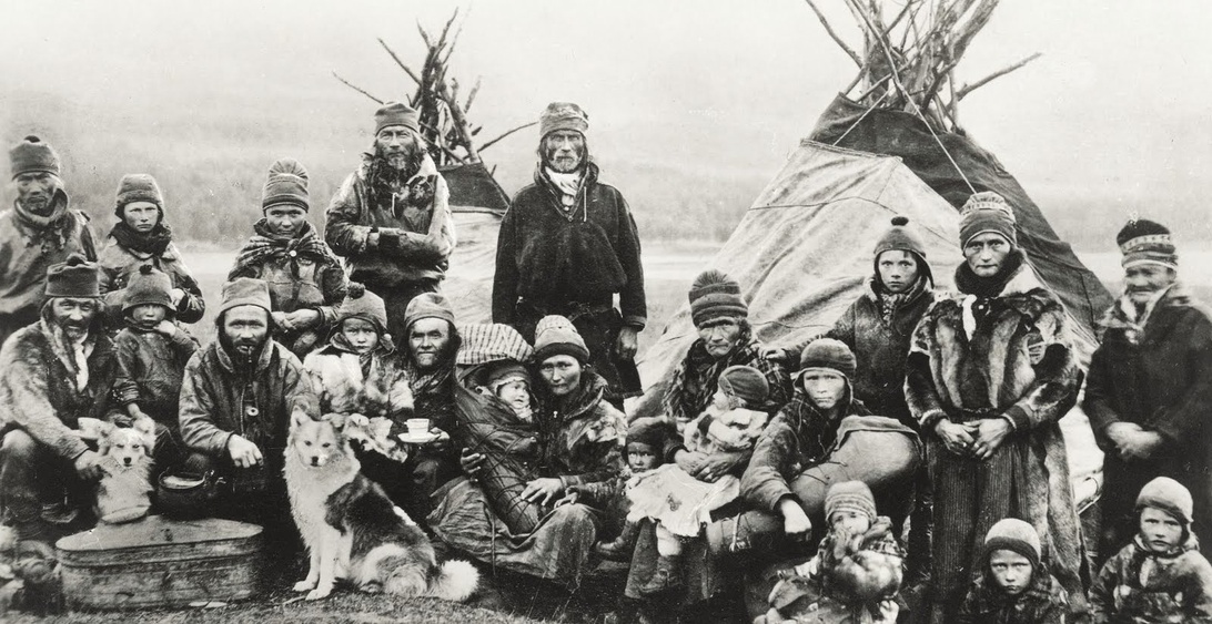 Historiskt fotografi av samer i traditionella kläder framför kåtor, vilket representerar en del av det rika och mångfacetterade nordiska kulturarvet.